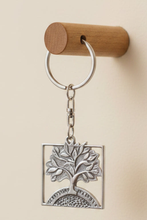 A hand holds a silver keychain on a dark backing card in a home setting. The keychain features a tree of life design inside a square frame.