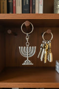 A hand holding a silver menorah keychain in its original clear plastic packaging card, which is branded 'Holy Holy Gifts'. The background is a cluttered outdoor or market setting with a woven chair and other items.