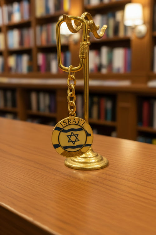 A gold-colored keychain with a small round medallion featuring the Israeli flag, held in a person's hand. The keychain is attached to a white cardboard backing inside a clear plastic sleeve, with a blurred background showing what appears to be a store or market setting.