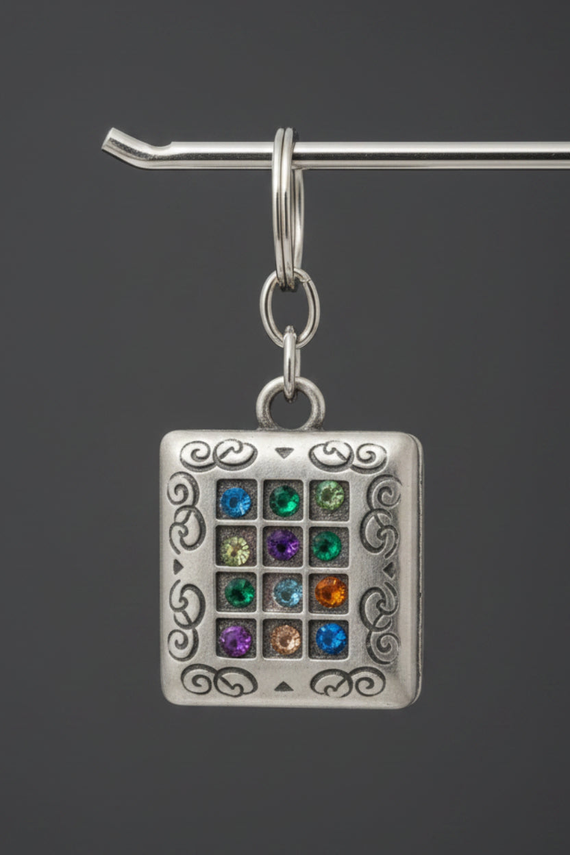 A hand holding a small silver-colored square pendant or charm wrapped in clear plastic. The pendant features an ornate engraved border and a central grid of colorful inlaid stones in green, blue, yellow, and red.